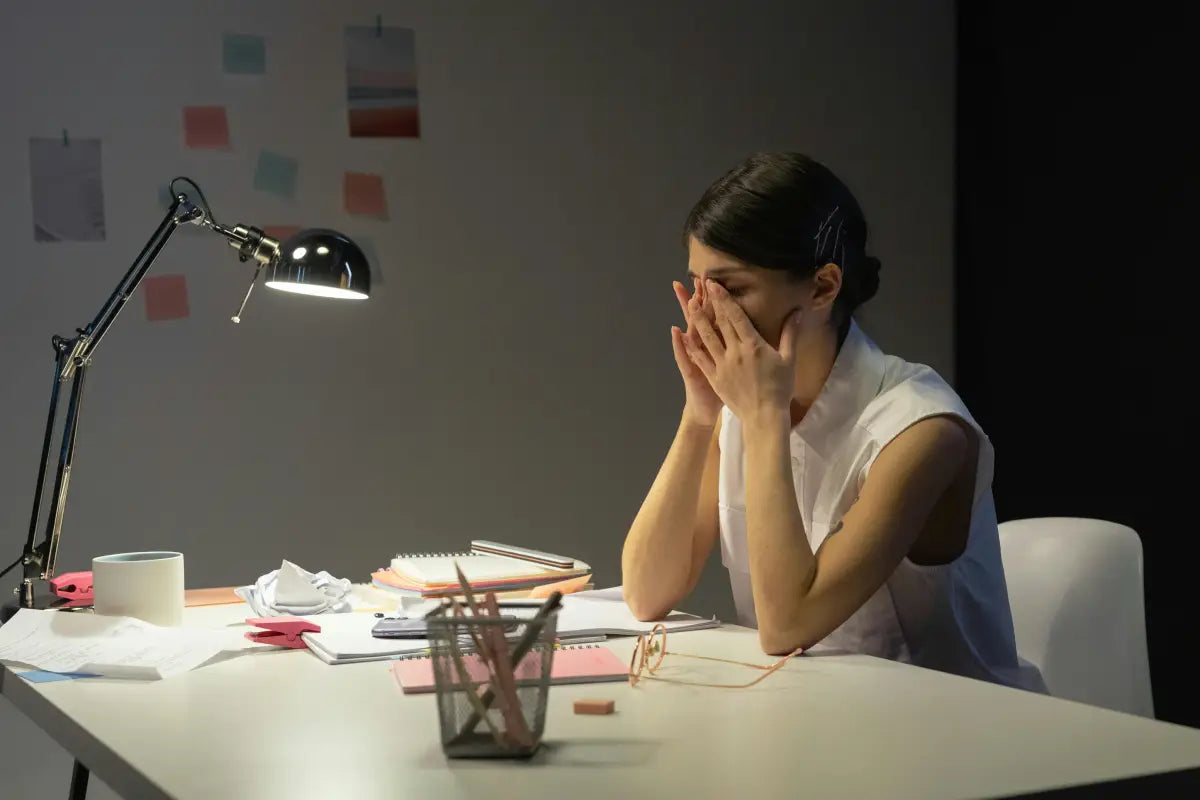 Eine Frau, die am Arbeitsplatz sitzt und gestresst wirkt. Sie fasst sich mit beiden Händen an die Augen.