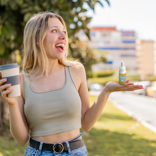 Eine Frau hält eine Flasche CBD-Öl in einer Hand, einen Becher Kaffee in der anderen.